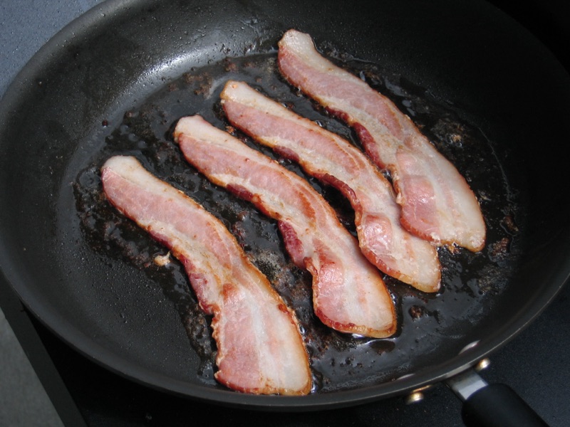 Four thick strips of bacon starting to render in a black skillet.