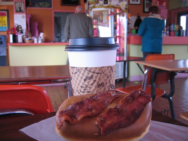 A maple-glazed doughnut topped with two strips of bacon next to a paper coffee cup.