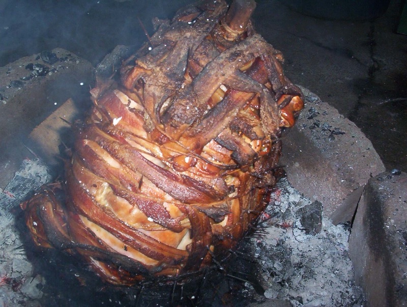 A turkey covered in a dark, crisp bacon lattice, standing upright over glowing coals.