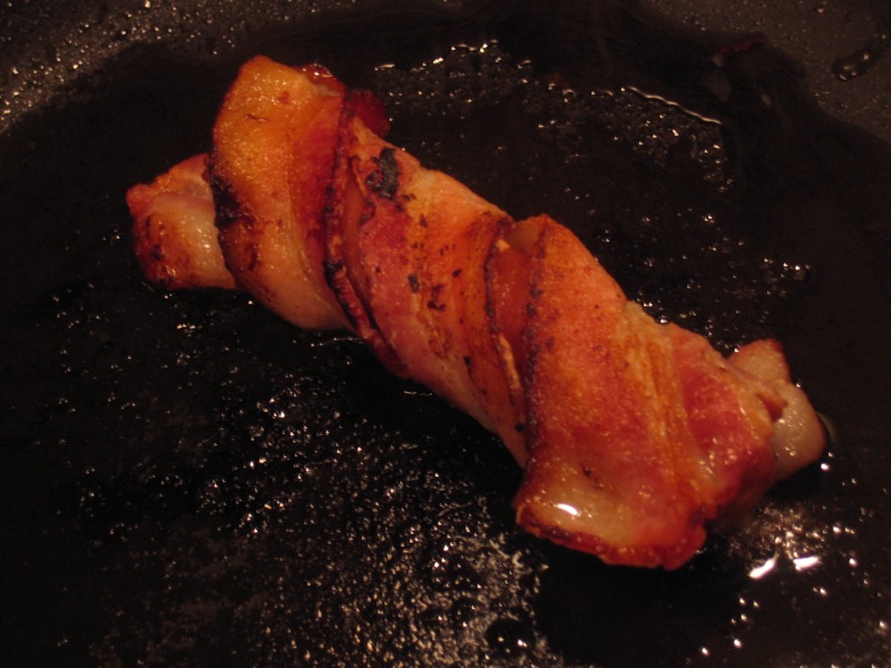 A sausage spiral-wrapped in bacon, browning in a dark skillet.