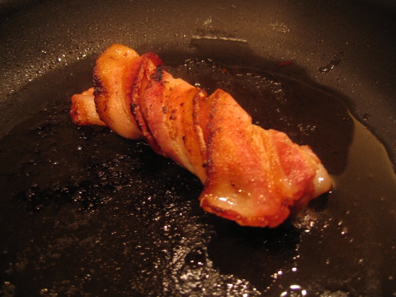 A bacon-wrapped sausage, crisp and blistered, in a skillet of rendered fat.
