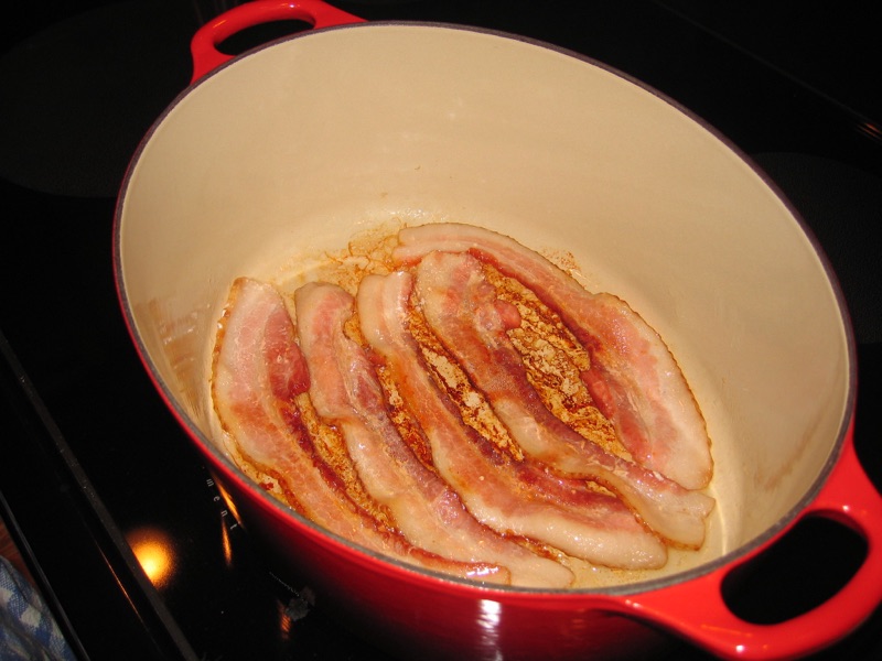 Bacon strips rendering in a red enameled Dutch oven.