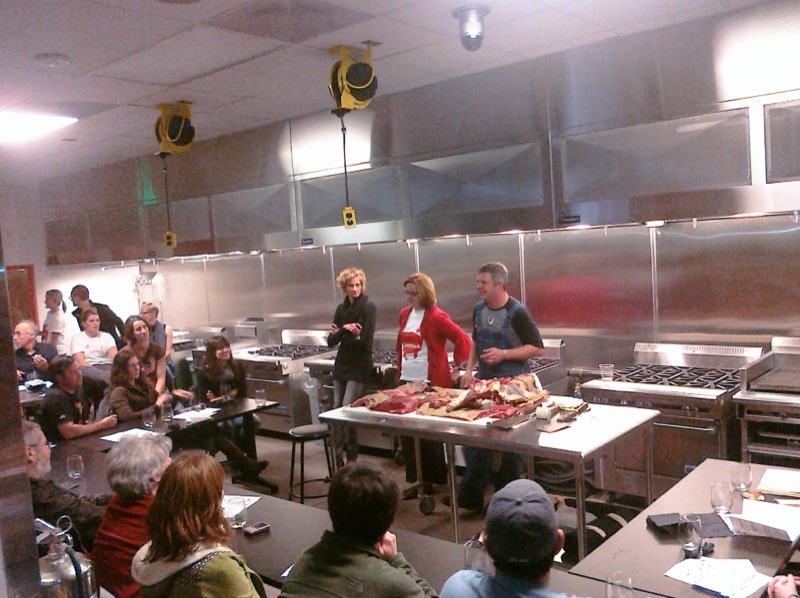 A teaching kitchen full of seated guests facing a steel table where butchered pork is laid out.