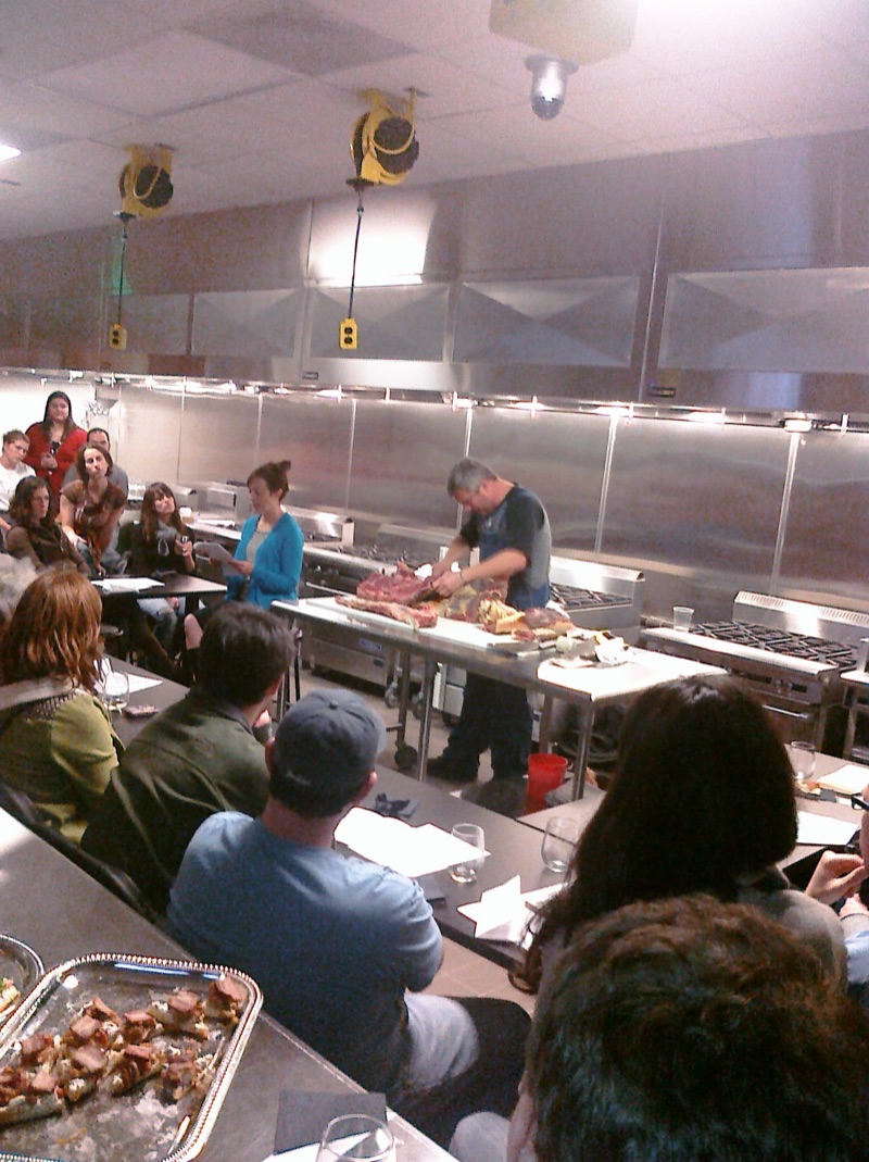 A butcher working at the steel table as guests lean in, a tray of cooked pork belly in the foreground.