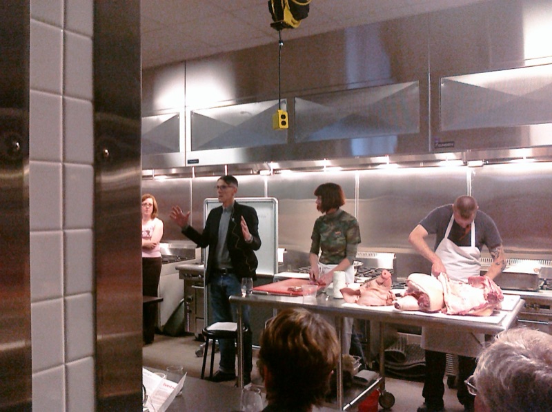 Two people in aprons working over a whole hog on a steel table while a third talks to the room.