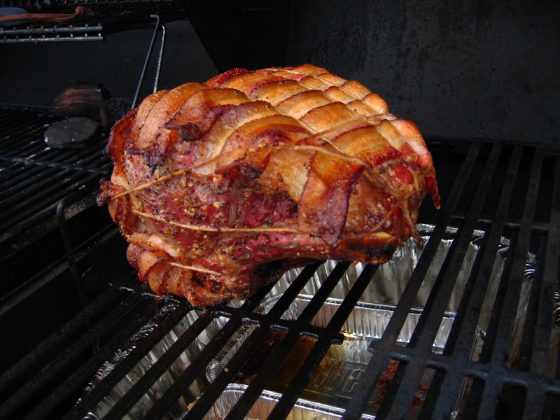 The bacon-wrapped roast browning on a dark grill grate over indirect charcoal.